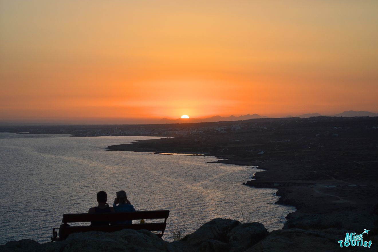 Two people sitting on a bench overlooking the ocean at sunset.