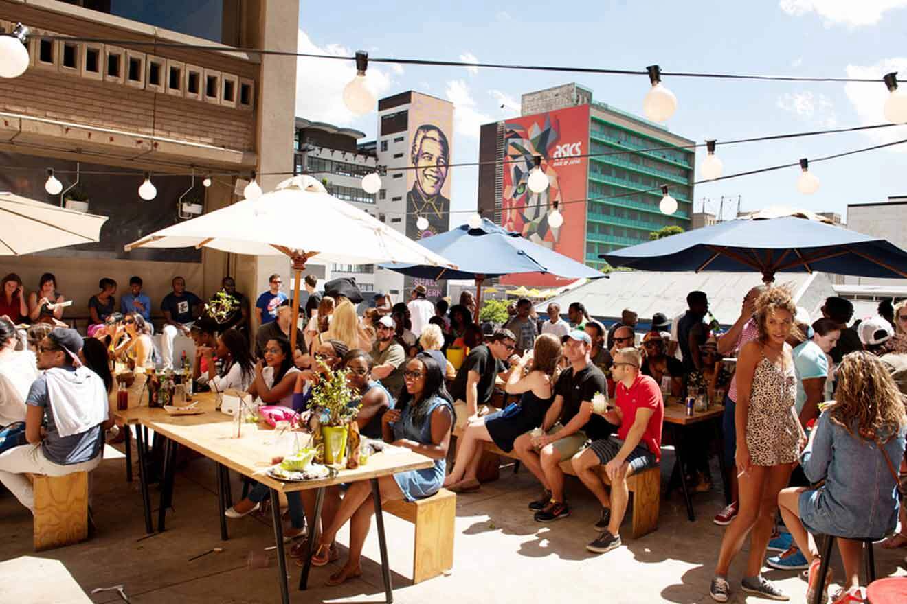 People sitting and standing around tables under umbrellas at an outdoor event during the daytime, with buildings and murals visible in the background.