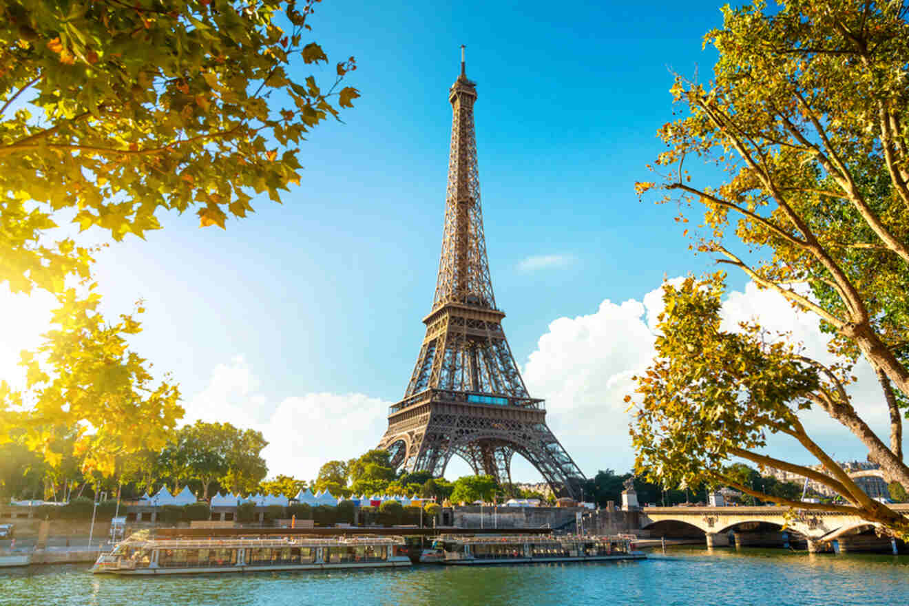 The Eiffel Tower in Paris viewed from the Seine River, surrounded by lush trees under a bright blue sky.