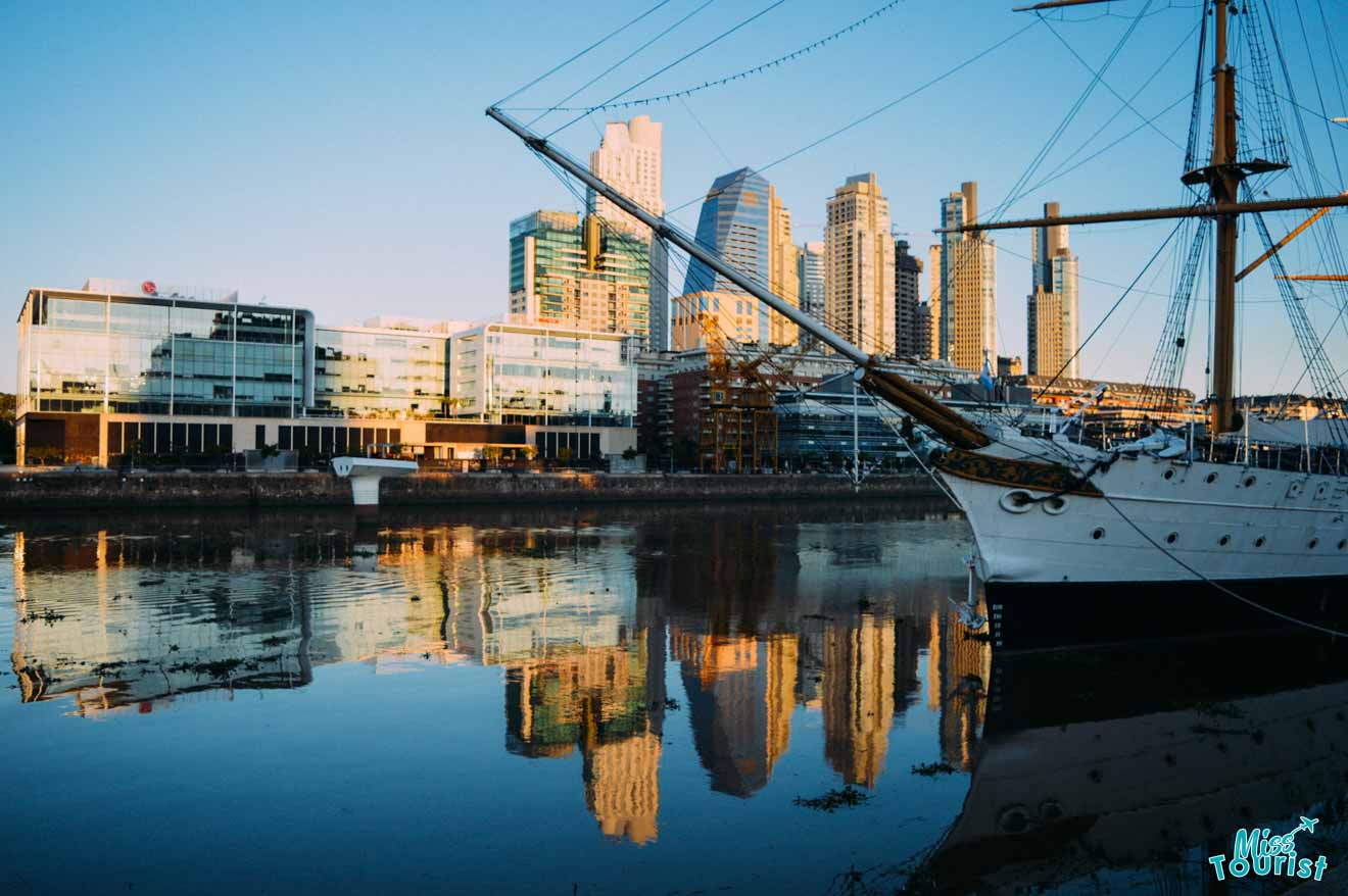 A serene waterfront scene at Puerto Madero in Buenos Aires, featuring the reflection of modern buildings and a historic ship, with the calm water providing a mirror image