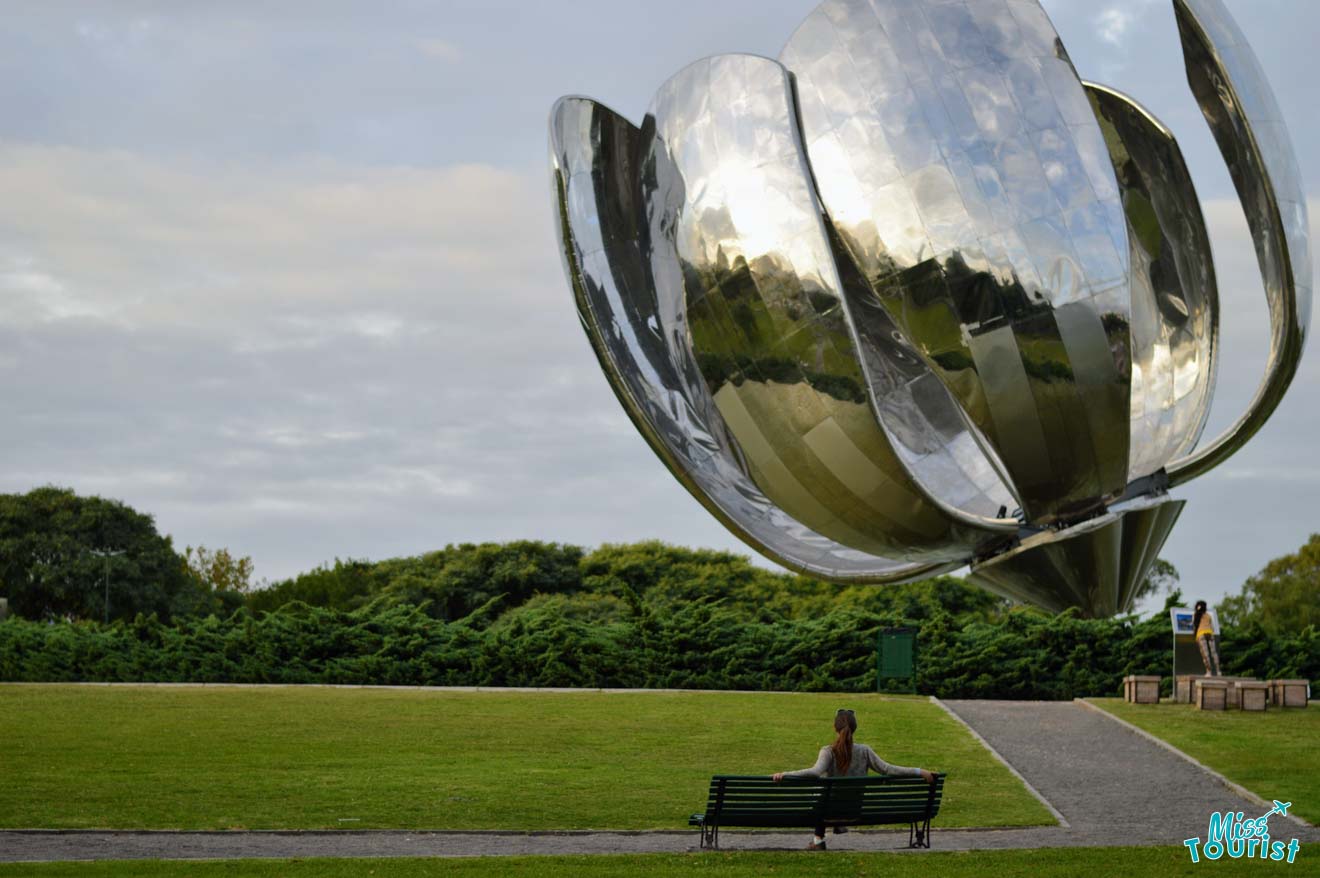 a woman sitting on a bench under a big metal flower-shaped sculpture
