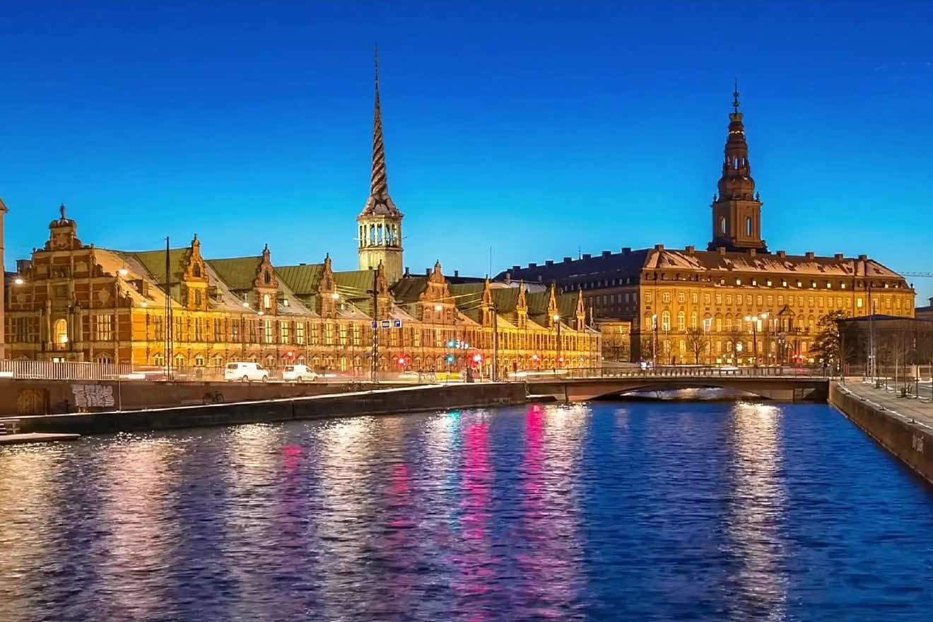 Nighttime view of the Børsen and Christiansborg Palace along the canal in Copenhagen, with reflections of lights on the water creating a picturesque scene