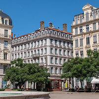 A classic European square with a multi-storied traditional hotel building featuring white facades and a greenery-filled plaza in the foreground