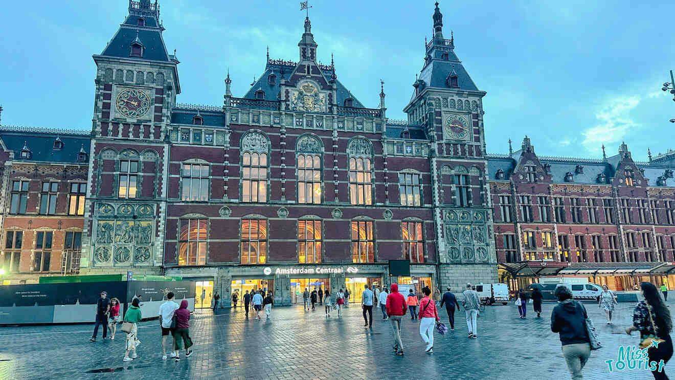 A view of Amsterdam Centraal station, an impressive Gothic-Renaissance building with intricate architectural details, illuminated by warm lights as people walk by in the evening.