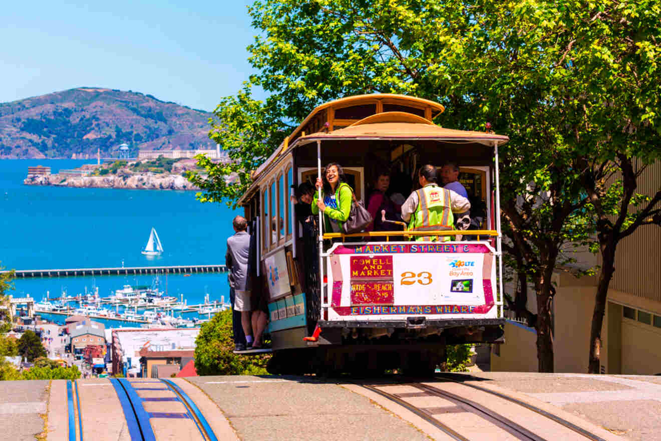 A traditional San Francisco cable car filled with passengers crests a hill, offering views of the blue bay and distant marinas, under the bright California sun.