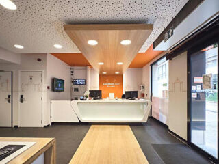 Hotel reception area with a sleek white desk, warm wood accents, and a welcoming orange backdrop