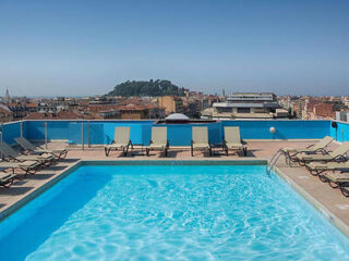 Rooftop swimming pool with sun loungers, overlooking a scenic view of Nice with the hill of Colline du Château in the distance, under a sunny sky