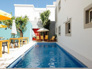 Narrow outdoor swimming pool flanked by white walls, with wooden poolside furniture and a vibrant yellow umbrella
