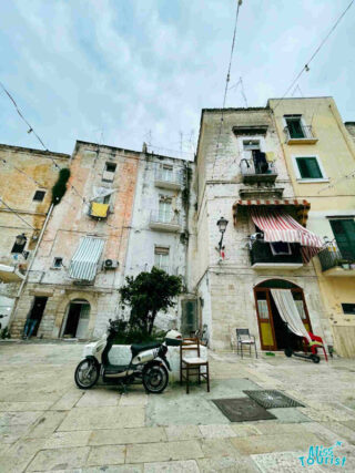A quiet, atmospheric square in Bari with worn historical buildings, a parked scooter, and a chair and table set on the cobbled stones, evoking a sense of local daily life.