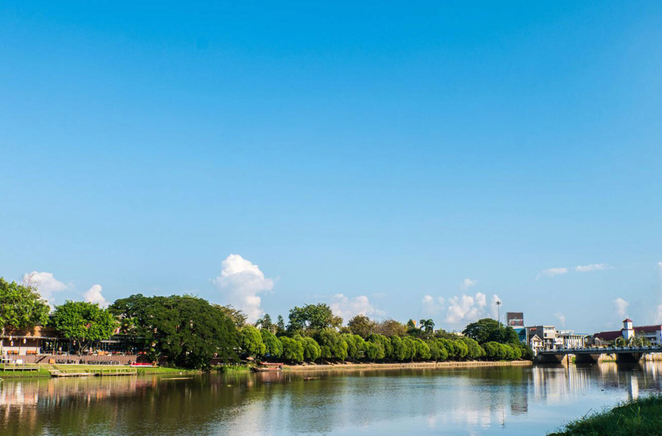 A serene riverside in Chiang Mai, with calm waters reflecting the lush greenery and clear blue skies, epitomizing a peaceful retreat.