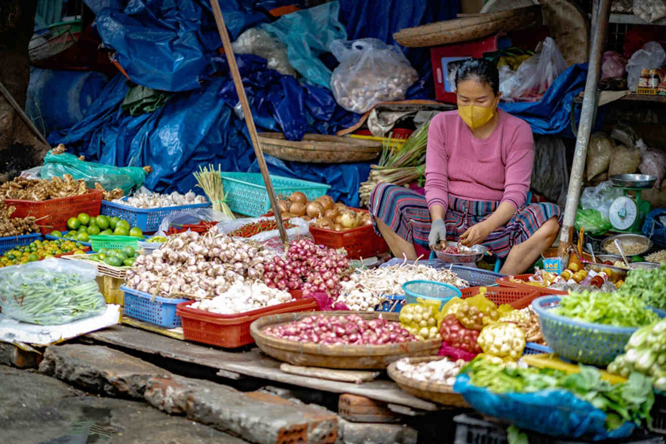 A local vendor in Bac My An Market, Da Nang, sits among an array of fresh produce including garlic, onions, and a variety of greens, reflecting the vibrant daily life and rich culinary culture in a traditional Vietnamese market setting