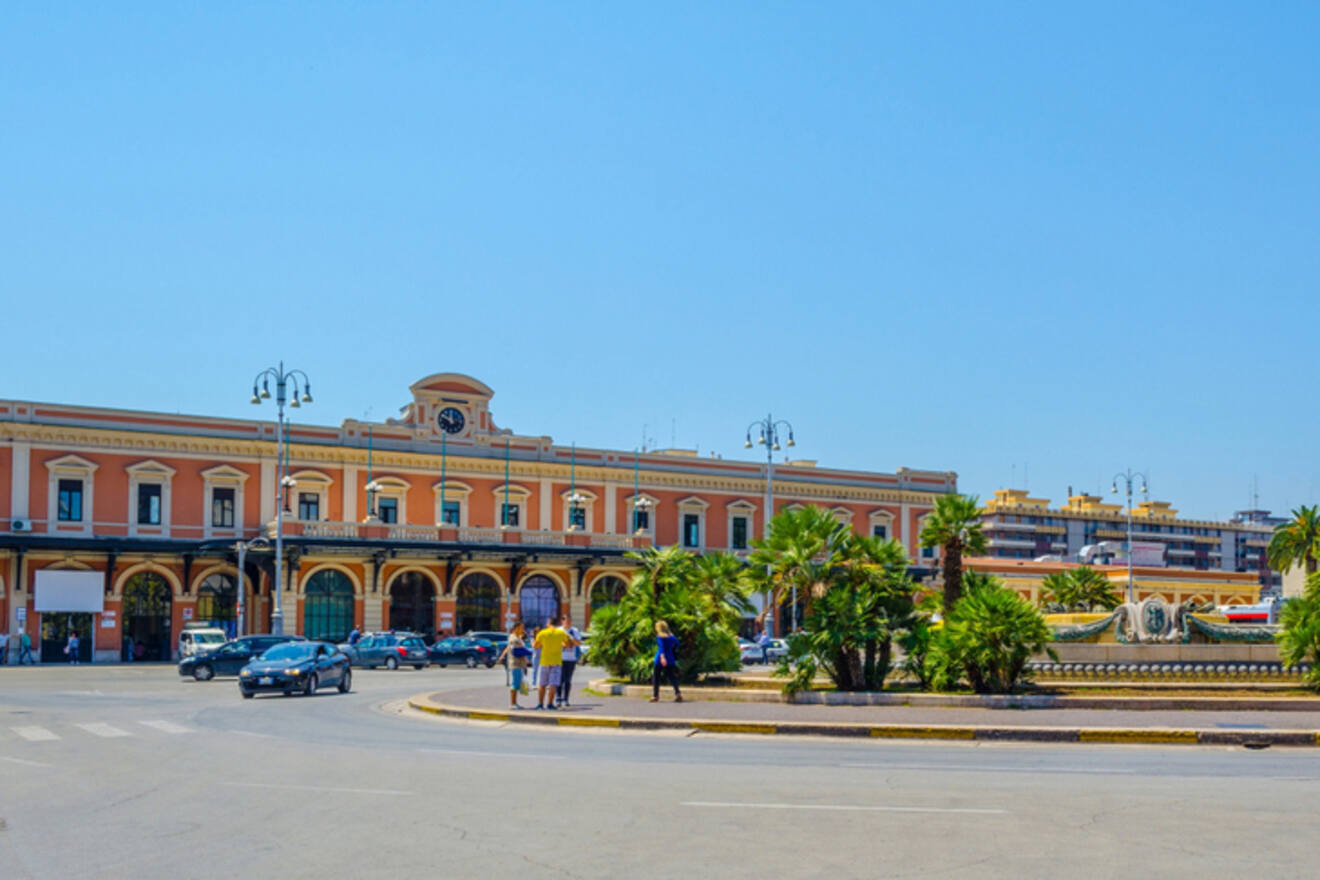 Panoramic view of a bustling square and the grand facade of Bari's train station with palm trees in the foreground