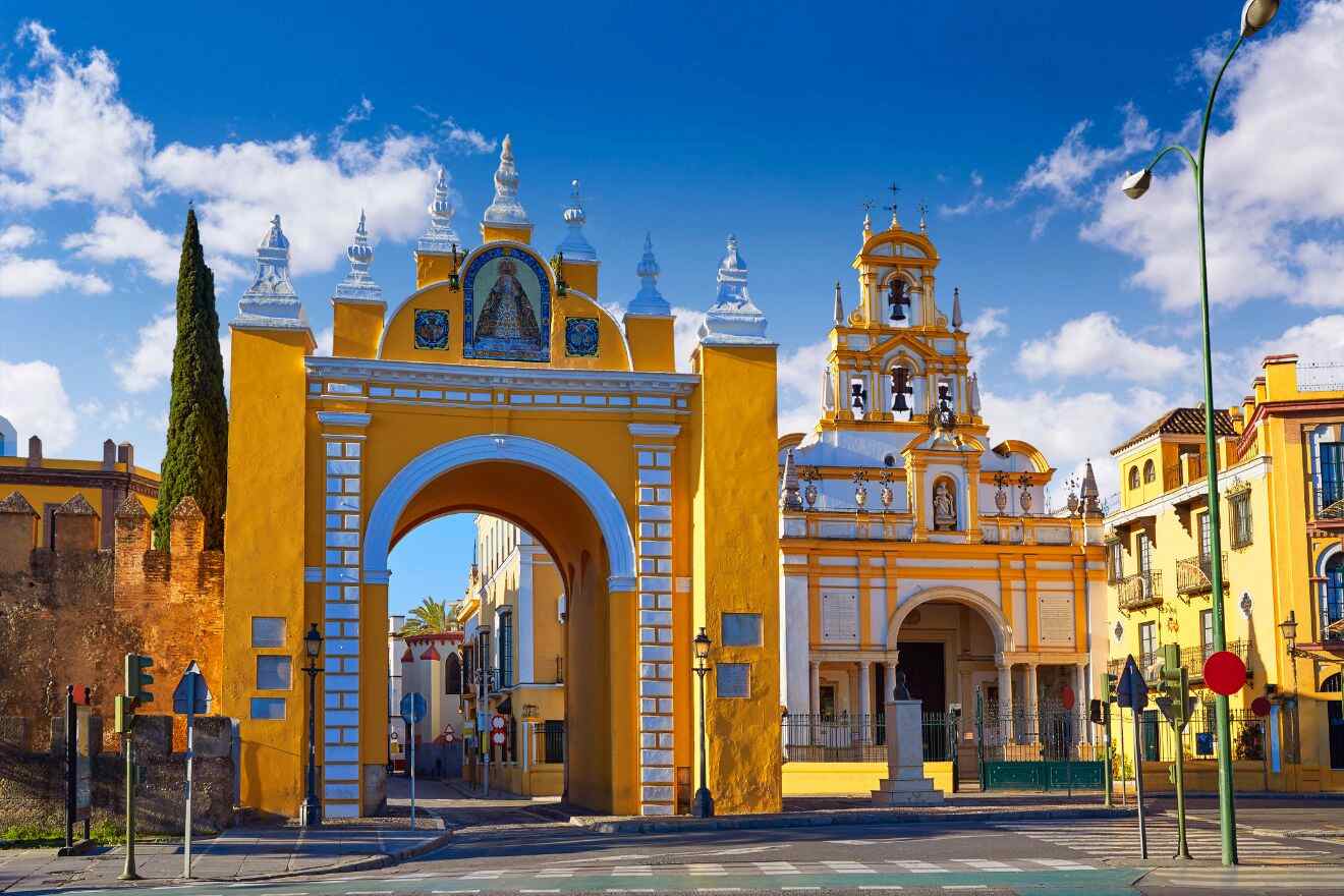 Vibrant yellow archway leading to the Basílica de la Macarena in Seville, Spain, under a bright blue sky. The Baroque church's intricate facade and ornate bell tower stand out against the historic city walls
