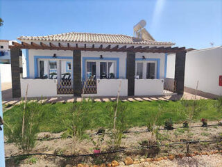 Small white guest house with blue trim and a terracotta tiled roof, set in a developing garden under a sunny sky