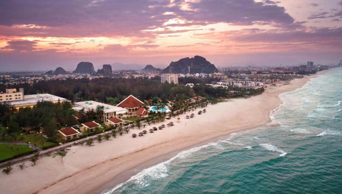 Panoramic dusk view of Non Nuoc Beach in Da Nang, showcasing the tranquil sea beside a stretch of luxury resorts and lush vegetation, with the distinctive Marble Mountains in the distance under a gradient sunset sky