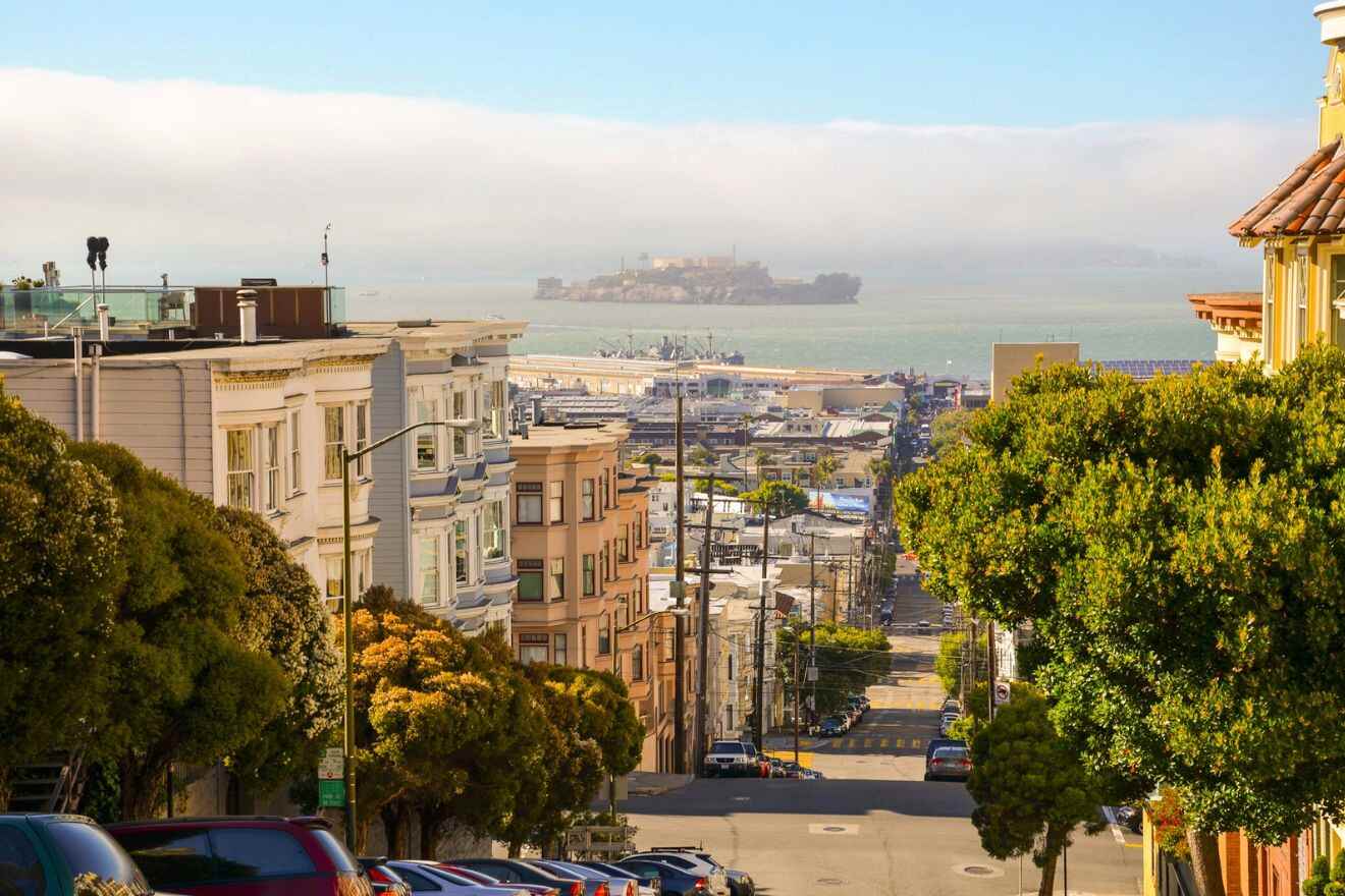 the steep descent of a San Francisco street lined with traditional architecture, framing the bay and marina in the background under a hazy sky.