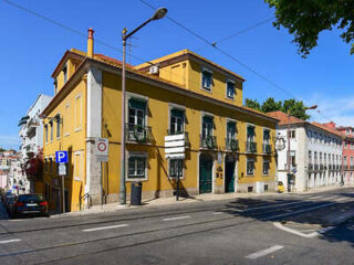 Sunny street view of a traditional yellow corner building with white windows in Lisbon, with cars parked alongside.