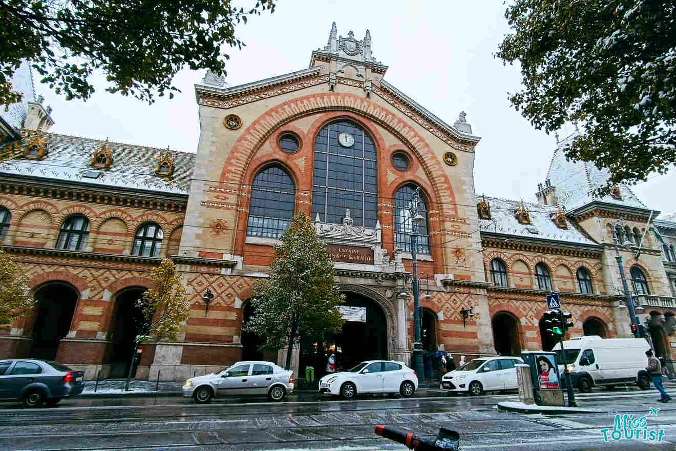 An ornate building of the Central Market Hall in Budapest with cars parked in front of it.