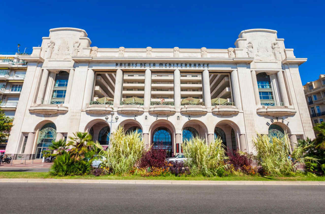 The iconic Palais de la Méditerranée hotel in Nice, showcasing its grandiose white Art Deco façade with bas-relief sculptures, surrounded by lush greenery and set against a clear blue sky