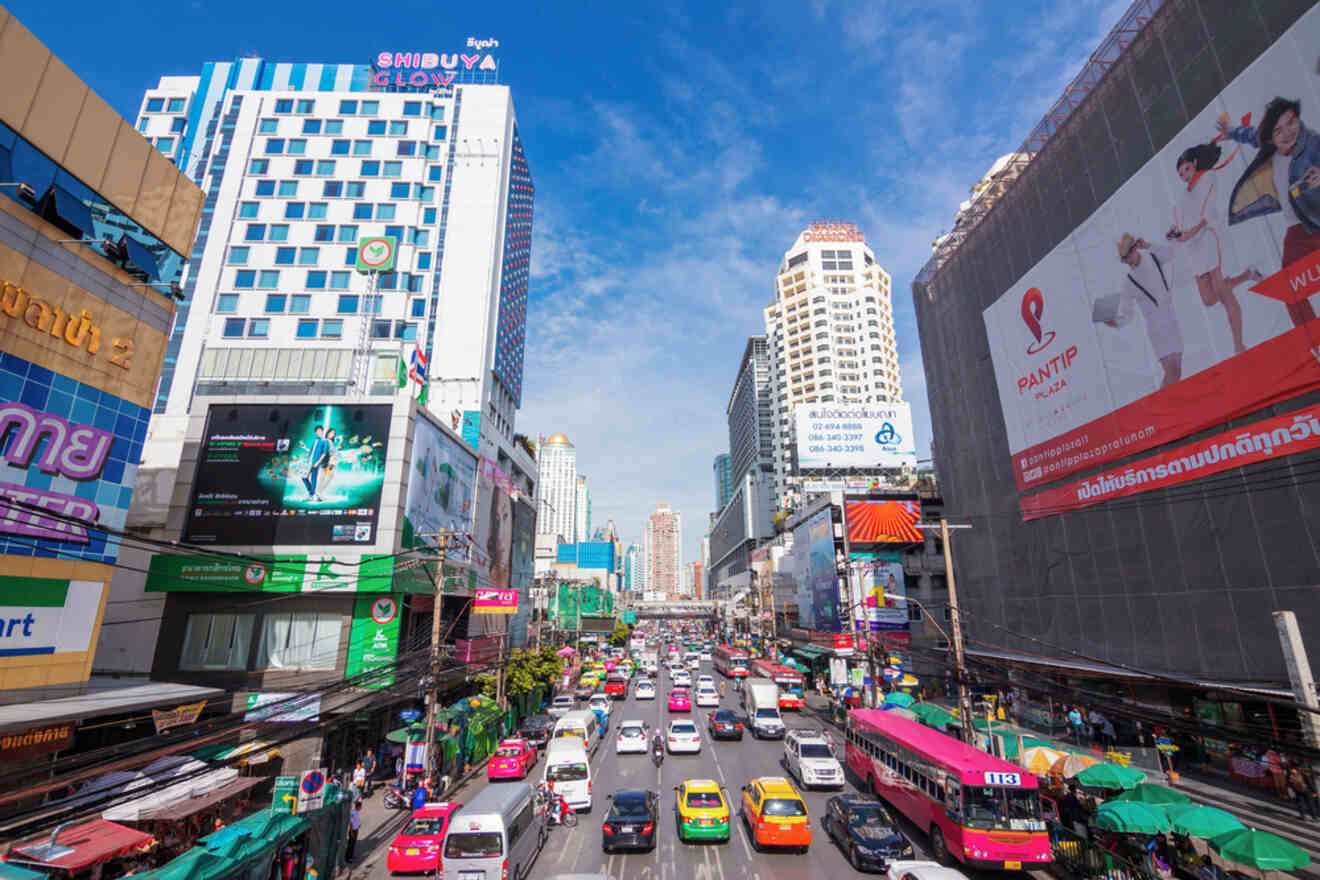Busy street scene in Pratunam area of Bangkok, with colorful taxis and tuk-tuks among modern high-rises under a clear blue sky