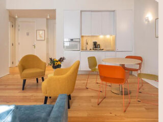 Bright and colorful open living space with a blue sofa, yellow chairs, an orange dining set, and a modern white kitchen in the background.