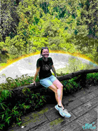 The writer of the post, Yulia Saf, smiling joyfully, seated on a wooden ledge with a faint rainbow in the mist behind her, surrounded by the lush greenery of a Chiang Mai forest.