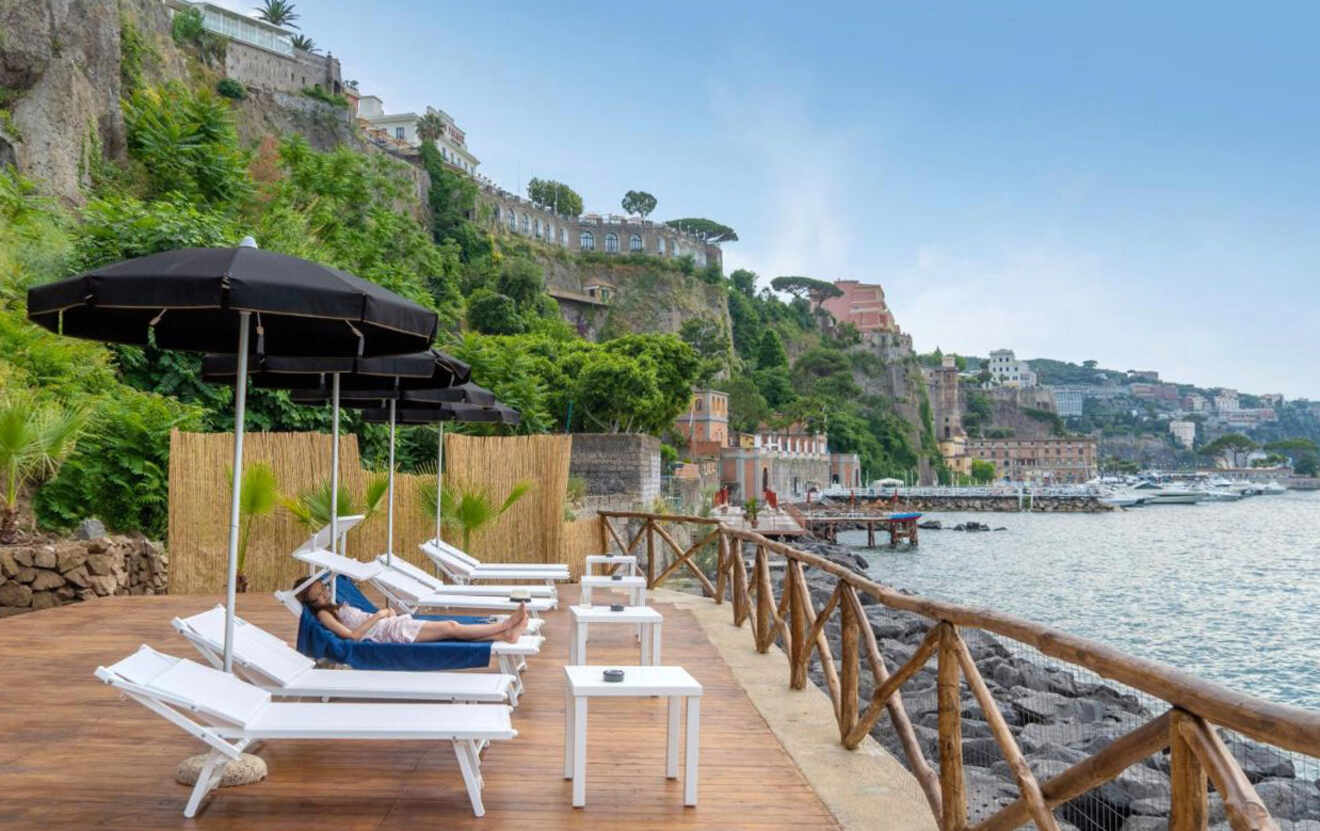 Waterfront relaxation area with white sun loungers and umbrellas, wooden deck, and a view of Sorrento's cliffside buildings in the background