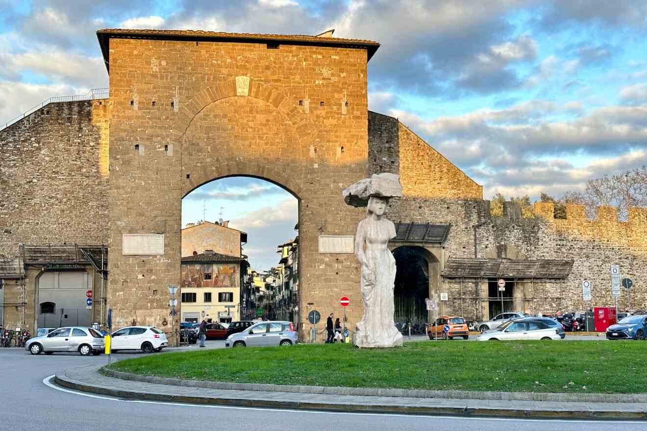 Porta San Niccolò, a historic city gate in Florence, Italy, stands against a clear blue sky, with surrounding traffic and a statue prominently displayed in the foreground