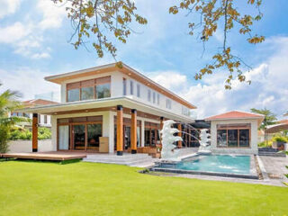 Modern two-story house with large windows, a green lawn, and a swimming pool featuring white decorative fountains under a partly cloudy sky.
