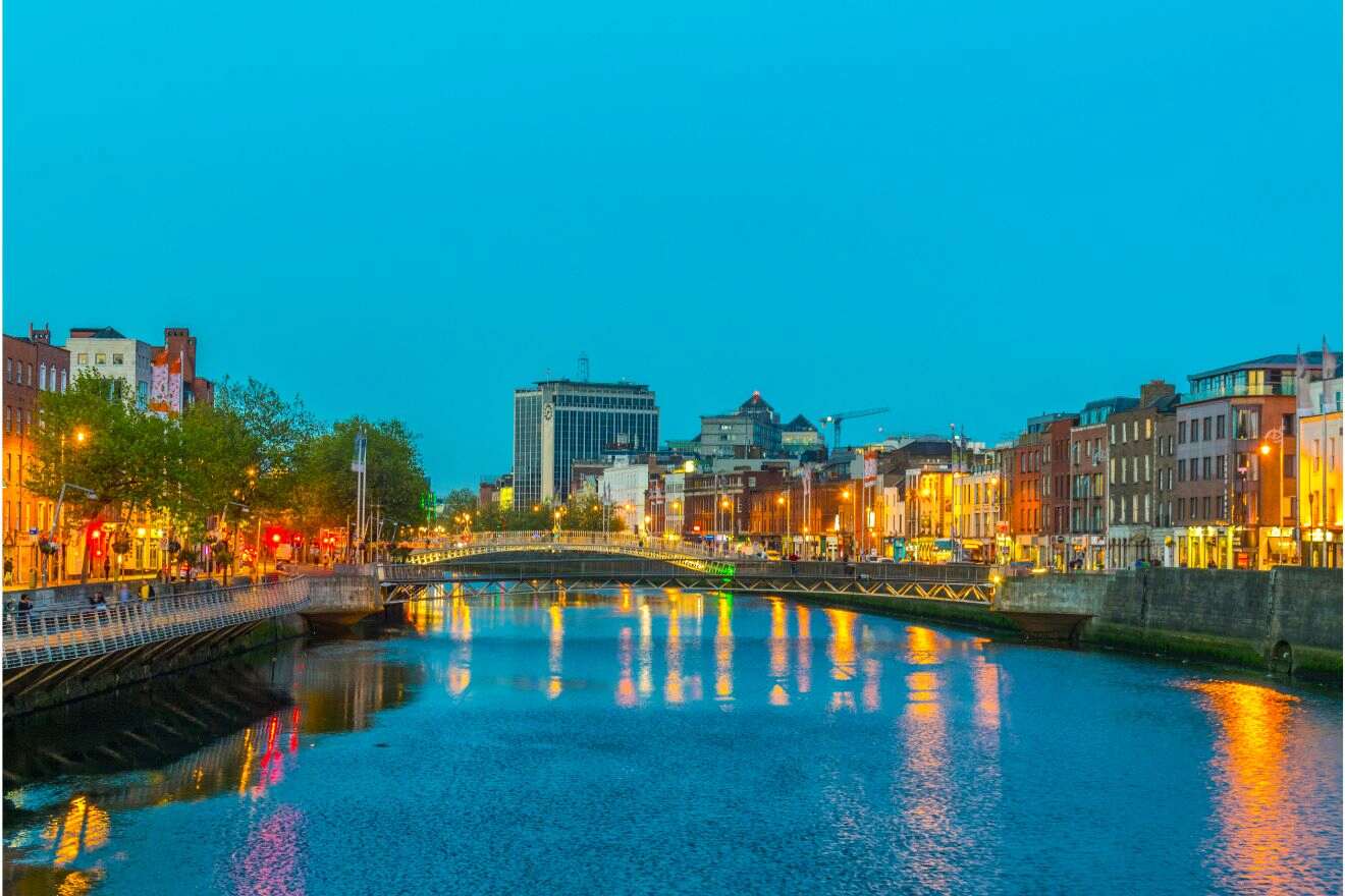 Twilight descends on Dublin as city lights reflect on the calm River Liffey, with the iconic Ha'penny Bridge connecting the illuminated streets lined with historical buildings
