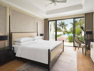 Modern hotel room with a large bed, two bedside tables, a desk, and glass doors opening to a patio with pool and palm trees outside.