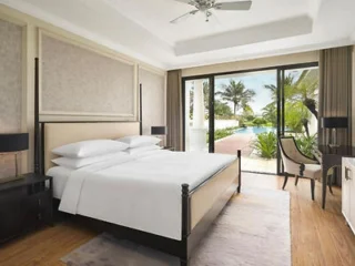 Modern hotel room with a large bed, two bedside tables, a desk, and glass doors opening to a patio with pool and palm trees outside.