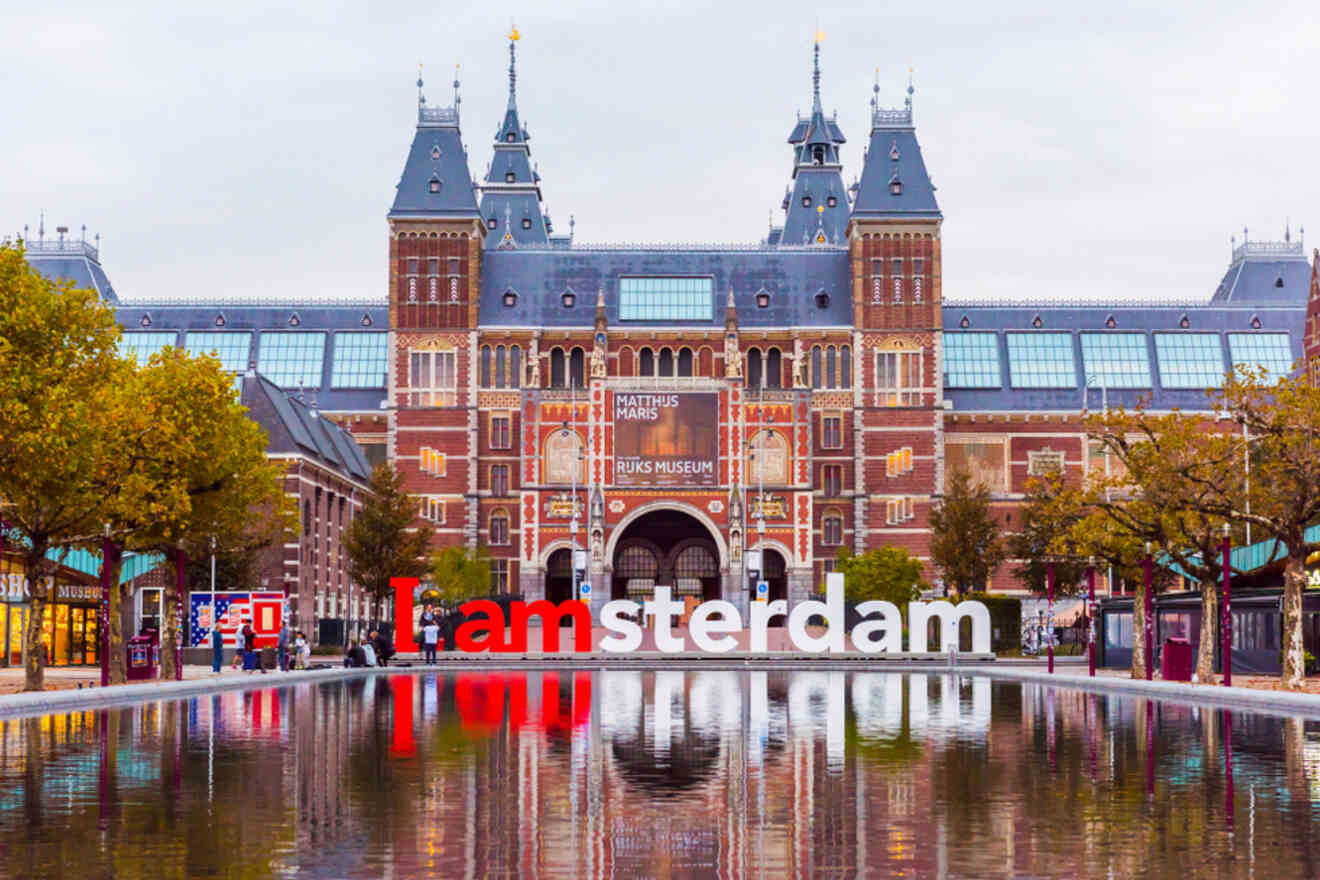 Iconic 'I amsterdam' sign in front of the historic Rijksmuseum in Amsterdam, reflecting in a pool of water, surrounded by trees and visitors