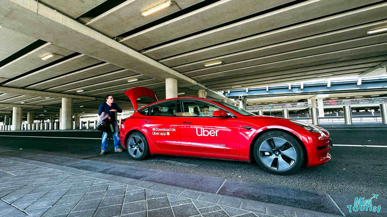 A bright red Uber car with a driver loading luggage into the trunk under the cover of a large, modern parking structure.