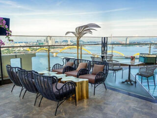 Rooftop patio with black wicker chairs, a gold table, and city views overlooking a river and a yellow bridge under a clear sky.