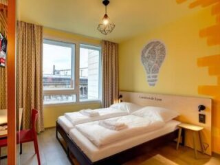 Hotel room with two single beds pushed together, white linens, a window with curtains, yellow walls, and a lightbulb-shaped wall graphic above the headboard.