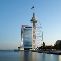 Modern waterfront building with a tall observation tower and a flag on top, next to a body of water under a clear sky at dusk.