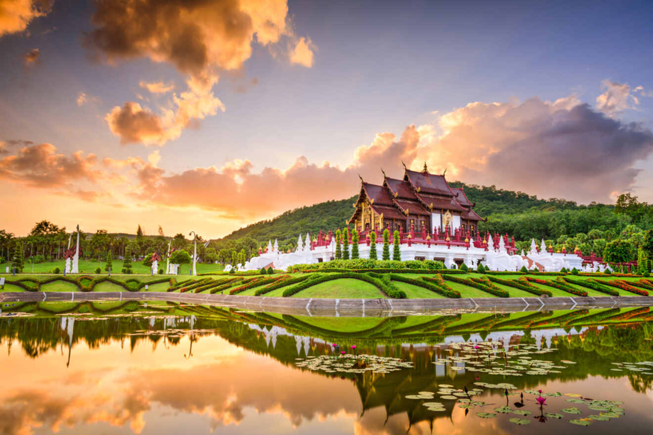 A majestic traditional Thai temple in Chiang Mai with intricate red and gold roofs, reflected in a tranquil pond amidst manicured green gardens under a vibrant sunset sky.