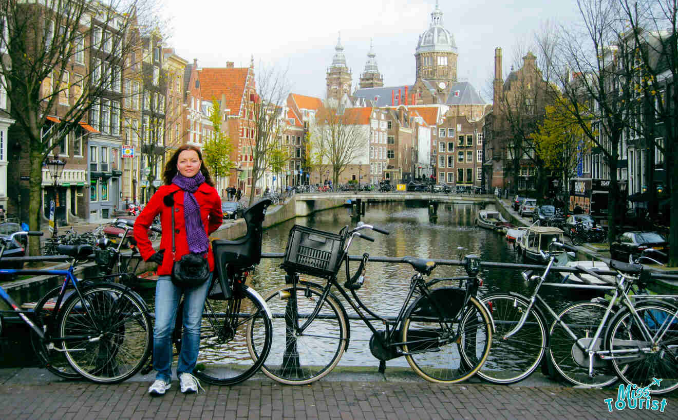 The writer of the post in a red coat and purple scarf stands next to parked bicycles on a bridge over a canal, with picturesque Dutch buildings and a church dome in the background.