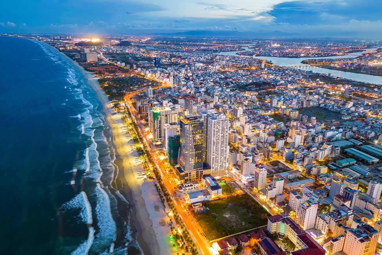 Aerial view of Da Nang city skyline at dusk with coastline, illuminated streets, and densely packed buildings