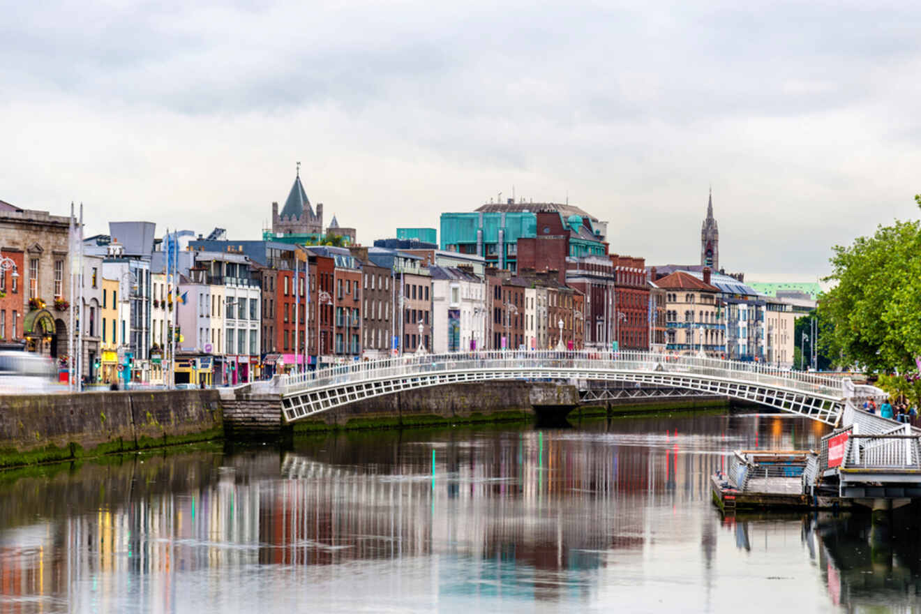 The Ha'penny Bridge arching over the River Liffey in Dublin, with a backdrop of varied city facades in muted colors, reflecting softly in the water on an overcast day.
