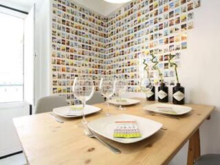 A dining table set for four with plates, glasses, and cutlery, in a room with two walls covered in a grid of small, colorful photo prints.