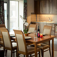 Well-appointed dining area with a wooden table set for a meal, cream chairs, and a kitchenette in the background