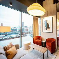 Chic corner of a city apartment with a large window offering a view of Manchester's skyline, furnished with a comfortable beige sofa, two velvety red armchairs, and a stylish yellow overhead lamp