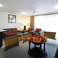 A spacious and contemporary living room with wicker furniture, plush red cushions, and a large glass dining table bearing a fruit bowl centerpiece