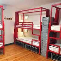 Hostel dormitory room featuring red metal bunk beds with white linens and personal lockers for storage.
