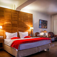 A hotel bedroom with a large bed with white bedding and red accents, wooden headboard and floor, ambient lighting