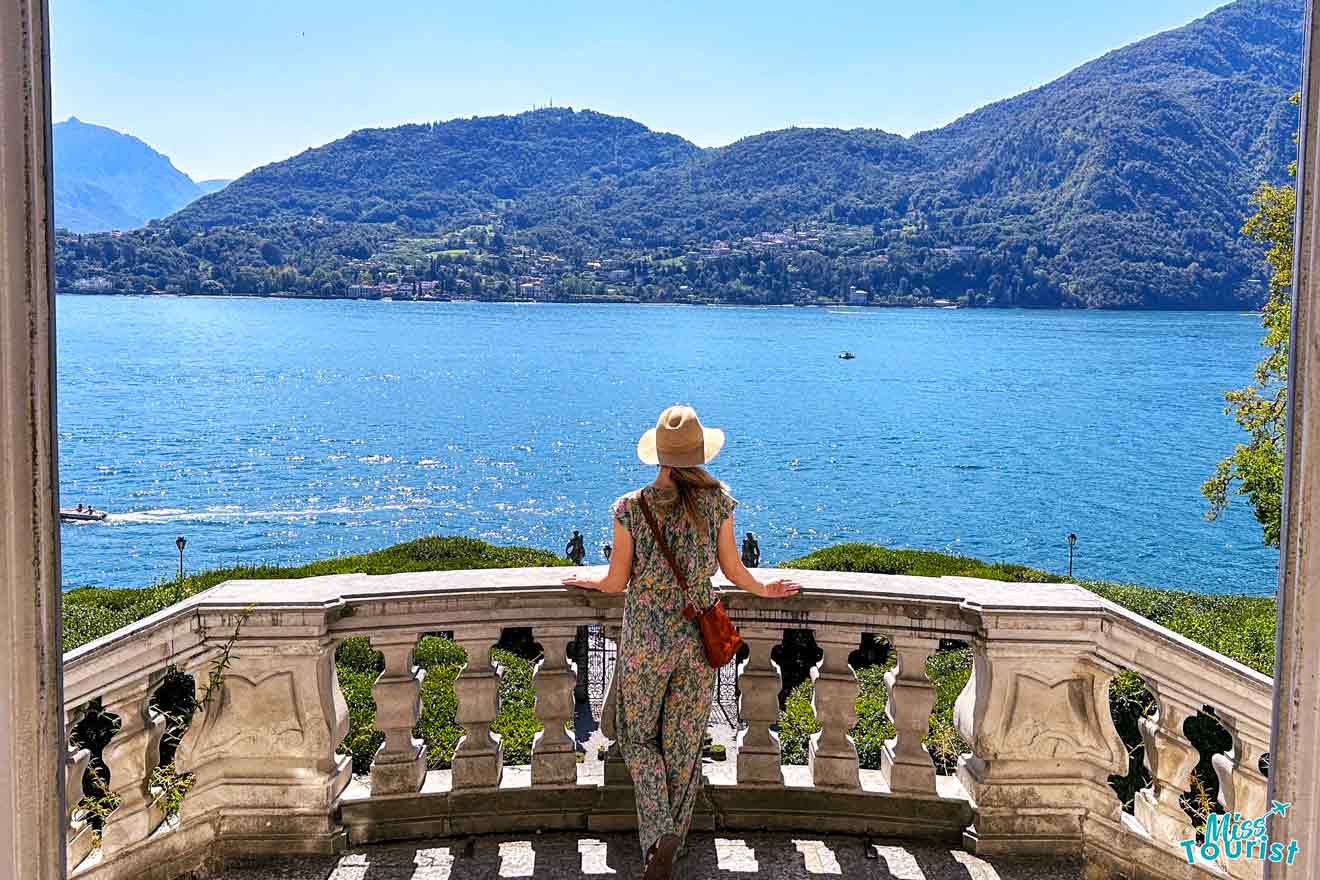 The writer of the post in a straw hat and floral dress stands on a balcony overlooking Lake Como, framed by lush green hills and a clear blue sky.