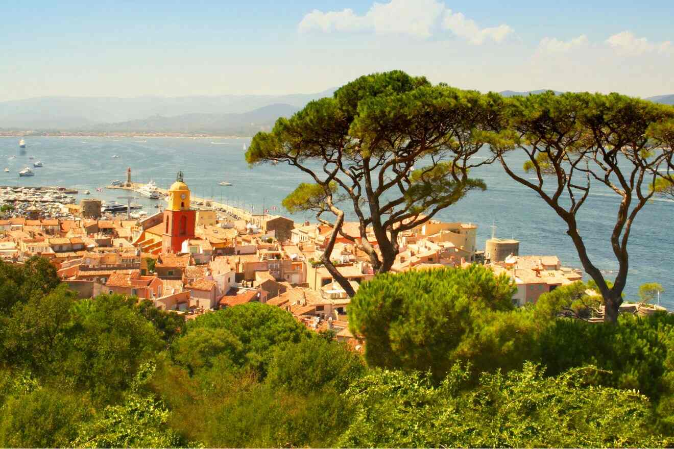Overlooking the terracotta rooftops of Saint-Tropez, this image captures the essence of the town with its iconic yellow bell tower, nestled among lush greenery and maritime pines, with the azure sea and moored boats in the background