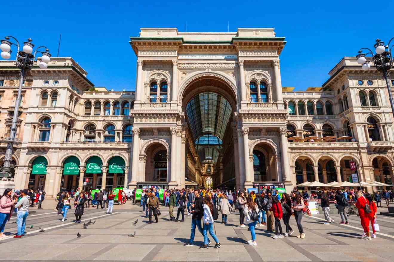 A busy public square in front of the ornate Galleria Vittorio Emanuele II in Milan, with pedestrians and pigeons amidst classical architecture under a clear sky